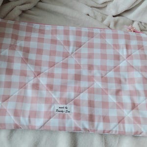 May include: A quilted, rectangular case with a pink and white gingham pattern. The case has a pink zipper and a small tag that reads "made by Pearly-Sue". The case is lying on a soft, white surface.