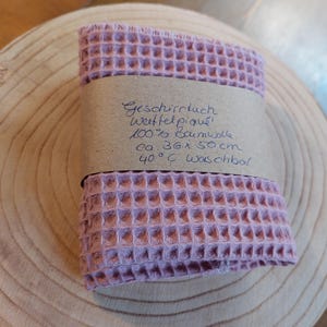 K&ouml;nnte beinhalten: Ein rosa Waffeltuch aus 100% Baumwolle. Das Tuch ist gefaltet und hat ein braunes Papier-Etikett mit dem Text "Geschirrtuch Waffelpique 100% Baumwolle ca. 36 x 50 cm 40&deg;C waschbar" befestigt.