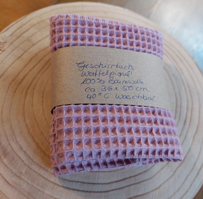 K&ouml;nnte beinhalten: Ein rosa Waffeltuch aus 100% Baumwolle. Das Tuch ist gefaltet und hat ein braunes Papier-Etikett mit dem Text "Geschirrtuch Waffelpique 100% Baumwolle ca. 36 x 50 cm 40&deg;C waschbar" befestigt.