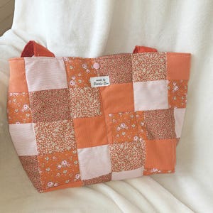 Könnte beinhalten: Eine Patchwork-Tragetasche in Orange-, Pfirsich- und Weißtönen. Die Tasche weist eine Vielzahl von Stoffen auf, darunter Blumenmuster und Streifenmuster. Die Griffe sind einfarbig orange, und ein kleines Etikett lautet "made by Pearla-Sue".