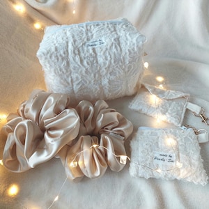 Könnte beinhalten: Eine Sammlung von Beauty-Accessoires, darunter zwei große Satin-Haargummis in hellem Champagner, eine strukturierte weiße Kosmetiktasche und zwei kleine weiße Spitzentaschen. Der Text "made by Pearly-Sue" ist auf den Artikeln sichtbar.