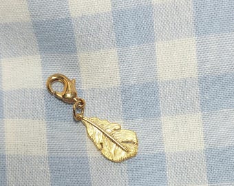 Könnte beinhalten: Ein goldfarbener Anhänger mit Federdesign. Der Anhänger ist an einem Verschluss befestigt, so dass er leicht an einer Halskette oder einem Armband befestigt werden kann. Der Hintergrund ist ein hellblau-weißes Karomuster.