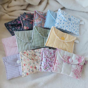 Può includere: Una collezione di piccole pochette in tessuto con vari motivi e colori. I disegni includono motivi floreali, a quadri e a righe. Ogni pochette ha una chiusura a bottone e un piccolo anello di tessuto.