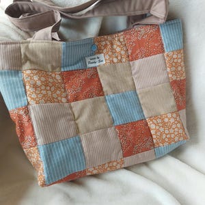 Op de afbeelding: Een patchwork tote bag met stoffen in oranje, blauw, beige en wit. De tas heeft een knoopsluiting en een schouderriem. Het label luidt "made by Pearly-Sue". De tas is gemaakt van verschillende stoffen met patronen.