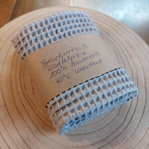 K&ouml;nnte beinhalten: Ein hellblaues Waffeltuch mit einem braunen Papierlabel, auf dem "Geschirrtuch Waffelpique 100% Baumwolle 40&deg;C waschbar" steht.