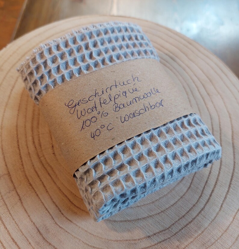 K&ouml;nnte beinhalten: Ein hellblaues Waffeltuch mit einem braunen Papierlabel, auf dem "Geschirrtuch Waffelpique 100% Baumwolle 40&deg;C waschbar" steht.