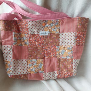 Könnte beinhalten: Eine Patchwork-Tragetasche mit rosa Trägern und einem Knopfverschluss. Die Tasche hat eine Vielzahl von Stoffen in Rosa-, Braun- und Weißtönen, darunter Punkte und Blumenmuster. Das Etikett lautet "made by Pearly-Sue."