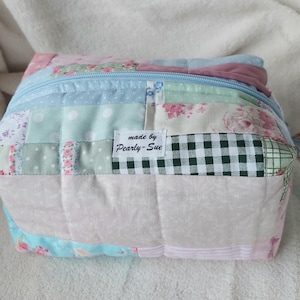 May include: A rectangular quilted pouch featuring a patchwork design of various pastel fabrics, including floral prints, polka dots, and a green and white gingham check. A light blue zip runs across the top, and a white label reads made by Pearly-Sue.