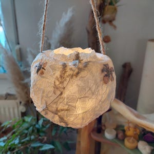 Könnte beinhalten: Eine weiße Papierlaterne mit getrockneten Blumen und Blättern im Inneren. Die Laterne hängt an einer Schnur und wird von innen beleuchtet, wodurch ein warmes Licht entsteht.
