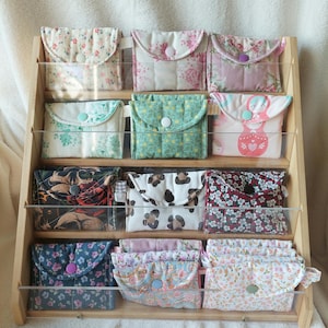 Può includere: Un espositore in legno contenente dodici piccole pochette in tessuto trapuntato con chiusura a scatto. Le pochette presentano vari motivi, tra cui stampe floreali, macchie di leopardo, un teschio e disegni di matrioske.