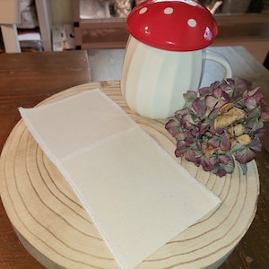 Könnte beinhalten: Eine weiße Keramiktasse mit einem roten und weißen Pilzdeckel. Die Tasse steht auf einem Holzuntersetzer mit einem weißen Stoffquadrat darauf. Ein Strauß getrockneter lila Blumen steht neben der Tasse.