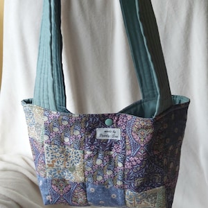 Könnte beinhalten: Eine Patchwork-Tragetasche mit einem Blumenmuster in Blau-, Lila- und Rosatönen. Die Tasche hat türkisfarbene Träger und ein türkisfarbenes Innenfutter. Ein kleines Etikett lautet "made by Pearly-Sue".