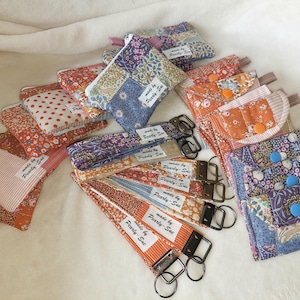 Peut inclure: Une collection d'articles en tissu faits à la main, comprenant des porte-clés, des pochettes et des bracelets, dans divers motifs floraux et patchwork. Les articles présentent une variété de couleurs, notamment orange, bleu et rouge. Chaque article porte une étiquette "made by Pearly-Sue".