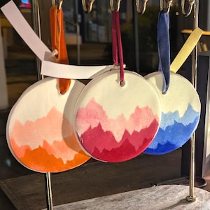 May include: A display of three round, white ceramic ornaments with colorful mountain range designs. The ornaments are painted with orange, red, and blue gradients. Each ornament hangs from a gold-colored metal stand with a marble base.