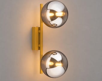 May include: A wall-mounted light fitting featuring a vertical yellow metal bar attached to a rectangular yellow base. Two spherical, semi-transparent smoked glass globes are mounted on the bar, each containing a glowing light bulb inside.