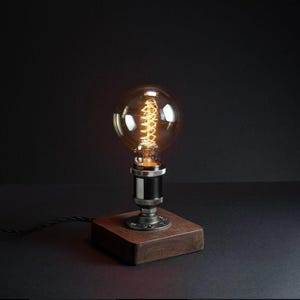 Op de afbeelding: Een tafellamp met een grote, ronde, heldere Edison-stijl gloeilamp met een zichtbare gloeiende spiraal. De lamp is gemonteerd op een vierkante voet van donker hout met een zwarte metalen pijpverbinding en een zwart snoer met stoffen bekleding.