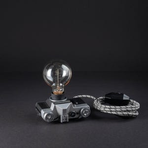 Könnte beinhalten: Eine Tischleuchte aus einem silber-schwarzen Vintage-Filmkameragehäuse. Eine große, klare, runde Glühbirne im Edison-Stil ist oben montiert. Die Leuchte verfügt über ein schwarz-weißes, stoffummanteltes Stromkabel mit einem schwarzen Zwischenschalter.