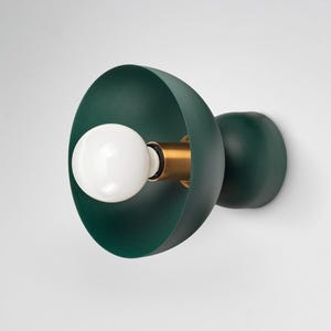 May include: A wall-mounted light fixture featuring a matte dark green bowl-shaped shade, a central brass-toned socket, and a round white globe light bulb. The fixture has a rounded dark green base that attaches directly to the wall surface.