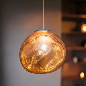 Peut inclure: Un luminaire suspendu avec un abat-jour sphérique en verre ambré. L'abat-jour a un effet texturé et fondu, et une ampoule est visible à l'intérieur. Le luminaire est suspendu par un fin cordon argenté.