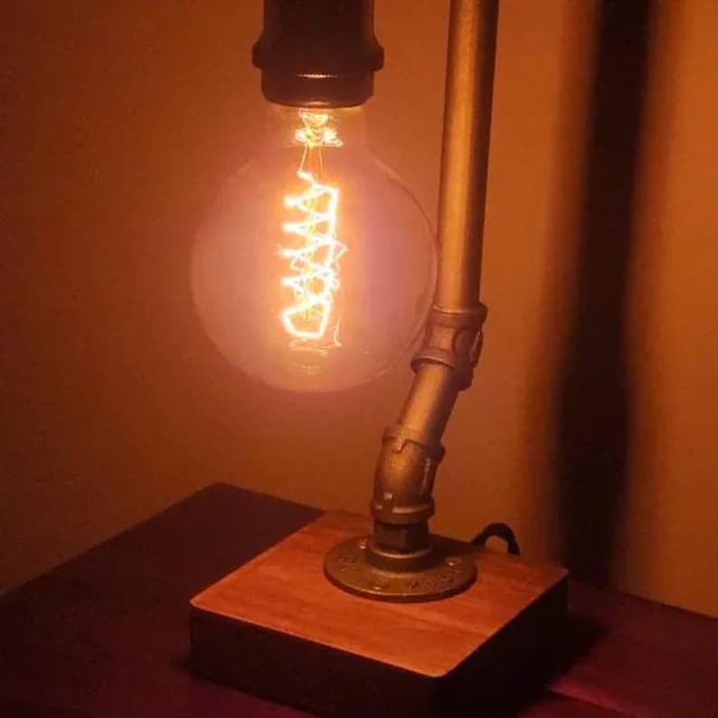 May include: A table lamp featuring a round, glowing Edison-style light bulb with a visible spiral filament. The lamp base is constructed from a square, polished wooden block, and the support arm is made of dark, metallic industrial pipe fittings.
