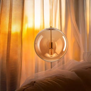 A spherical, amber-coloured pendant light fitting. The glass globe houses a visible light bulb and is suspended by a thin, gold-toned rod. The background features sheer curtains and warm lighting.