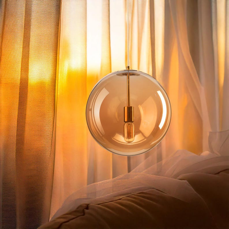 A spherical, amber-coloured pendant light fitting. The glass globe houses a visible light bulb and is suspended by a thin, gold-toned rod. The background features sheer curtains and warm lighting.