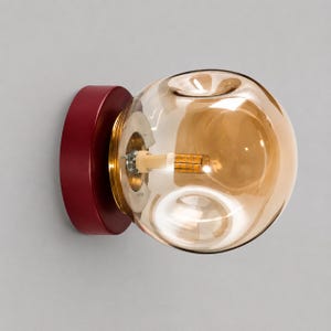 May include: A wall-mounted light fixture featuring a round, deep red base and a large, spherical, amber-tinted glass shade. The interior reveals a central metallic gold-toned mounting plate and a small, cylindrical LED light bulb component.