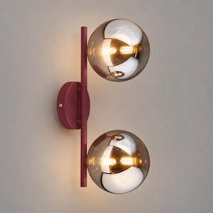 May include: A wall sconce featuring a vertical matte burgundy metal rod attached to a circular wall mount. Two spherical, amber-tinted glass globes are mounted on the rod, each containing a warm, glowing light source that reflects the surroundings.