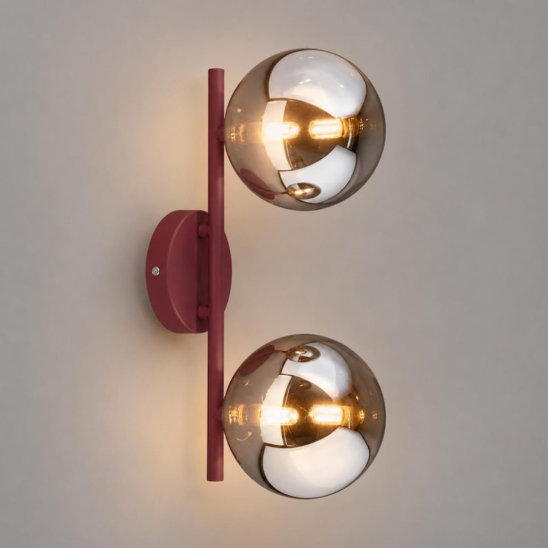 May include: A wall sconce featuring a vertical matte burgundy metal rod attached to a circular wall mount. Two spherical, amber-tinted glass globes are mounted on the rod, each containing a warm, glowing light source that reflects the surroundings.