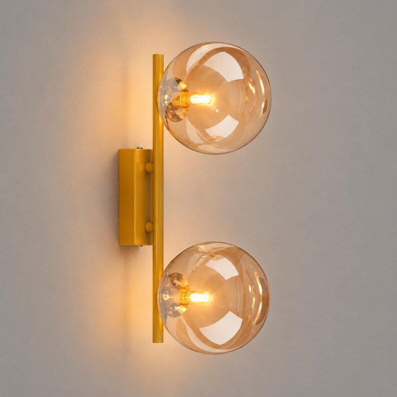 May include: A wall-mounted light fixture featuring a vertical yellow metal bar attached to a rectangular base. Two spherical, amber-tinted glass globes are mounted on the bar, each containing a glowing light bulb that emits a warm, soft light.
