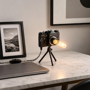 Può includere: Una lampada a forma di macchina fotografica Zenit nera, con base a treppiede e lampadina Edison accesa. È posizionata su una superficie di marmo bianco, accanto a un laptop e a una foto incorniciata.