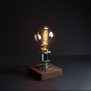 Puede incluir: Una lámpara de mesa de estilo vintage con una bombilla Edison. La lámpara presenta una gran bombilla de vidrio transparente con un filamento visible, montada sobre una base metálica y un bloque de madera. La lámpara está diseñada para una mesa o escritorio.