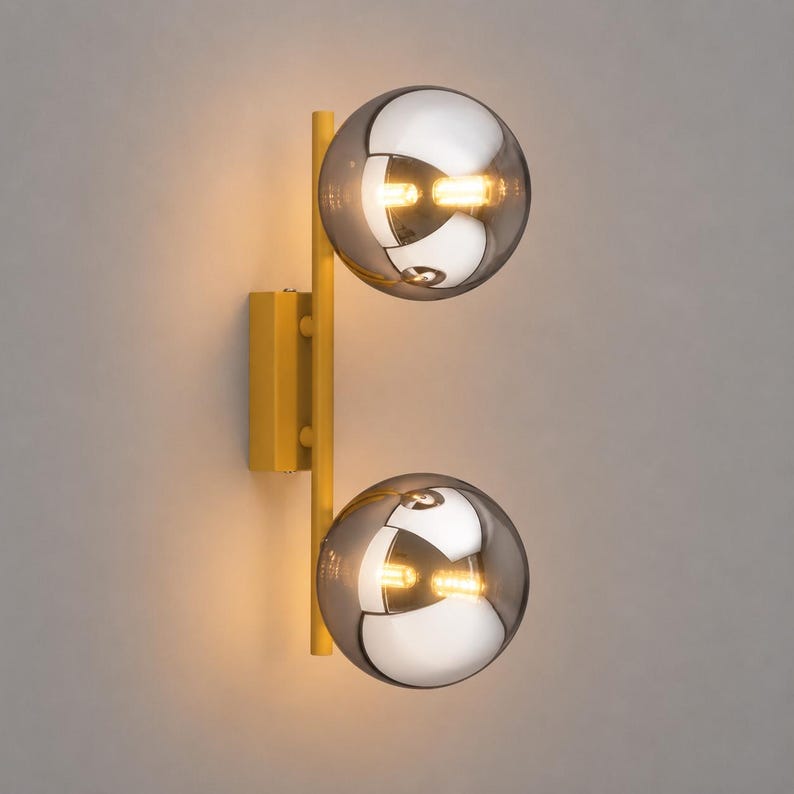 May include: A wall-mounted light fixture featuring a vertical yellow metal bar attached to a rectangular yellow base. Two spherical, semi-transparent smoked glass globes are mounted on the bar, each containing a glowing light bulb inside.