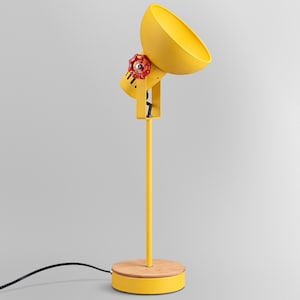 Może przedstawiać: Jasnożółta lampa biurkowa z kloszem w kształcie misy, pionowym stojakiem i okrągłą drewnianą podstawą. Lampa posiada czerwone pokrętło regulacyjne w kształcie zaworu z boku klosza oraz czarny przewód zasilający wychodzący z podstawy.