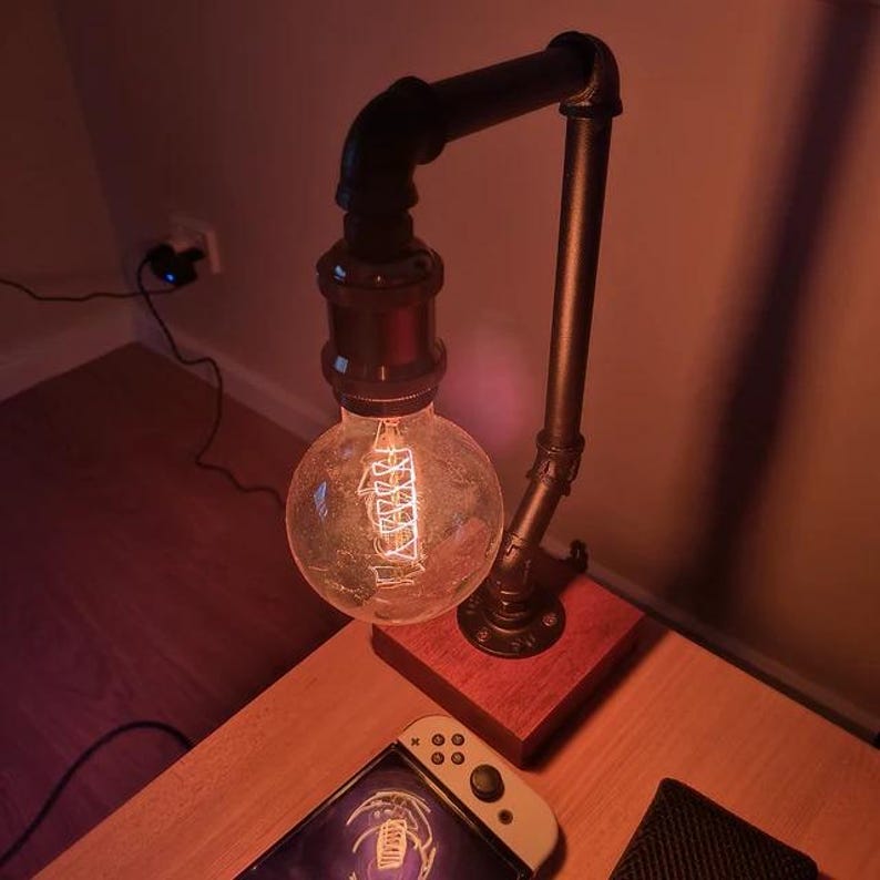 May include: A table lamp constructed from dark gray industrial metal piping, featuring a round Edison-style light bulb with a visible glowing filament. The lamp is mounted on a square wooden base and emits a warm, orange-toned light.