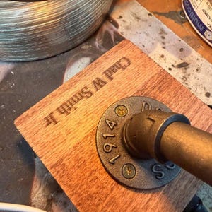 May include: A square wooden base with the engraved text Chad W Smith Jr, featuring a dark gray circular metal pipe flange attached with two gold-colored screws. A metallic pipe extends from the center of the flange, which is embossed with the number 1914.