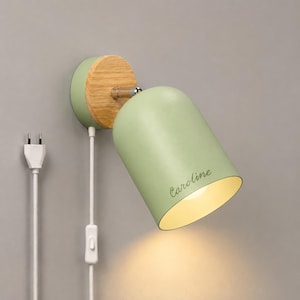 Op de afbeelding: Een wandlamp met een saliegroene, koepelvormige kap waarop de naam Caroline in cursief schrift is gedrukt. Het armatuur is voorzien van een ronde voet van licht hout, een zilverkleurig metalen scharnier en een wit snoer met een schakelaar.
