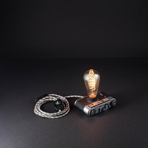 Könnte beinhalten: Eine Kamera im Vintage-Stil, die als Tischlampe umfunktioniert wurde, mit einem silber-schwarzen Metallgehäuse und einer leuchtenden Edison-Glühbirne an der Oberseite. Ein stoffummanteltes Stromkabel mit schwarzem Schalter ist beigefügt.