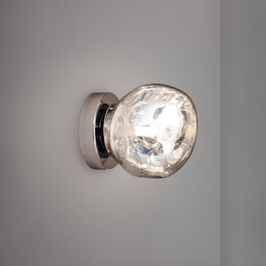 Puede incluir: Un aplique de pared con una base circular de cromo pulido y una pantalla de vidrio de forma orgánica. El vidrio tiene una superficie texturizada y ondulada que difunde la luz, creando un brillo luminoso y reflectante desde el interior.