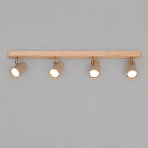 May include: A linear ceiling light fixture featuring a long, light-toned wooden base mounted with four cylindrical, beige-colored spotlights. Each spotlight is attached to the wooden bar by a small, metallic gold-toned swivel joint.