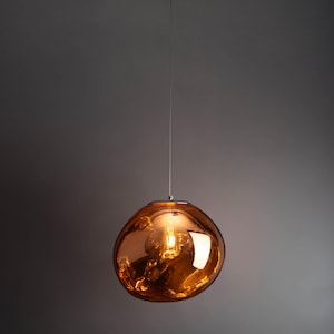 Könnte beinhalten: Eine moderne, kugelförmige Pendelleuchte mit einem warmen, bernsteinfarbenen Glasschirm. Die Leuchte hängt an einem dünnen Silberdraht, und die Glühbirne im Inneren ist sichtbar. Das Design ist minimalistisch und elegant, passend für verschiedene Einrichtungsstile.