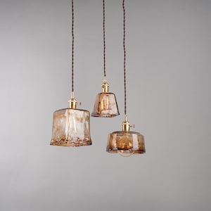 May include: Three pendant lights with amber-colored glass shades and brass fixtures hang from brown cords. The shades have a textured appearance, and the bulbs are visible inside. The lights are suspended against a neutral gray background.
