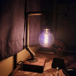 May include: A table lamp constructed from dark grey metal plumbing pipes and fittings, mounted on a square wooden base. The lamp features a large, round, clear glass Edison-style light bulb with a visible glowing filament, emitting a warm light.