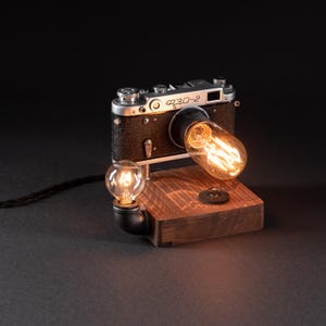 Könnte beinhalten: Eine Tischleuchte aus einem alten FED-2 Kameragehäuse auf einem rechteckigen Holzsockel. Die Leuchte verfügt über eine längliche Edison-Glühbirne am Objektivanschluss und eine kleinere runde Glühbirne an einem schwarzen Metallrohranschluss.