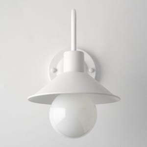 May include: A matte white wall sconce featuring a circular mounting plate, a vertical arm, and a wide conical shade. A large, spherical, frosted white light bulb is centered beneath the shade, providing a clean and uniform appearance.