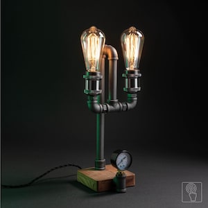 Puede incluir: Lámpara de mesa de estilo industrial con dos bombillas Edison. La lámpara está construida con tuberías y accesorios de metal negro, montados sobre una base de madera. Se adjunta un manómetro. Tiene una estética vintage y steampunk.