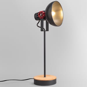 Może przedstawiać: Czarna metalowa lampa biurkowa ze złotym wnętrzem klosza, wyposażona w charakterystyczne czerwone pokrętło regulacyjne w kształcie zaworu. Lampa jest osadzona na cienkim pionowym słupku przymocowanym do okrągłej podstawy z jasnym drewnianym blatem.