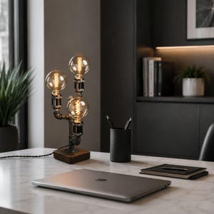 May include: An industrial-style desk lamp with three Edison light bulbs connected by metal pipes, set on a wooden base. The lamp sits on a marble-effect surface, alongside a laptop, pen holder, and notebook. The design has a vintage feel.