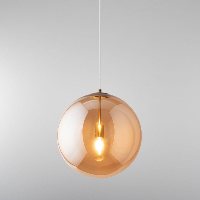 A spherical pendant light with an amber glass shade. The light bulb inside is lit, emitting a warm glow. The fixture is suspended by a thin, white cord, offering a modern, minimalist aesthetic. The shade's diameter is approximately 30 cm.