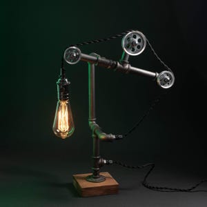 May include: Industrial-style desk lamp with a vintage Edison bulb. The lamp features a metal pipe frame, pulley system, and a wooden base. The bulb is a warm amber color, and the lamp has a black cord.